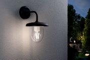 Schwarze Außen-Wandleuchte im Laternenstil mit transparentem Glas und sichtbarem Leuchtmittel an grauer Hauswand, warm leuchtend, Perspektive: leicht seitliche Frontansicht