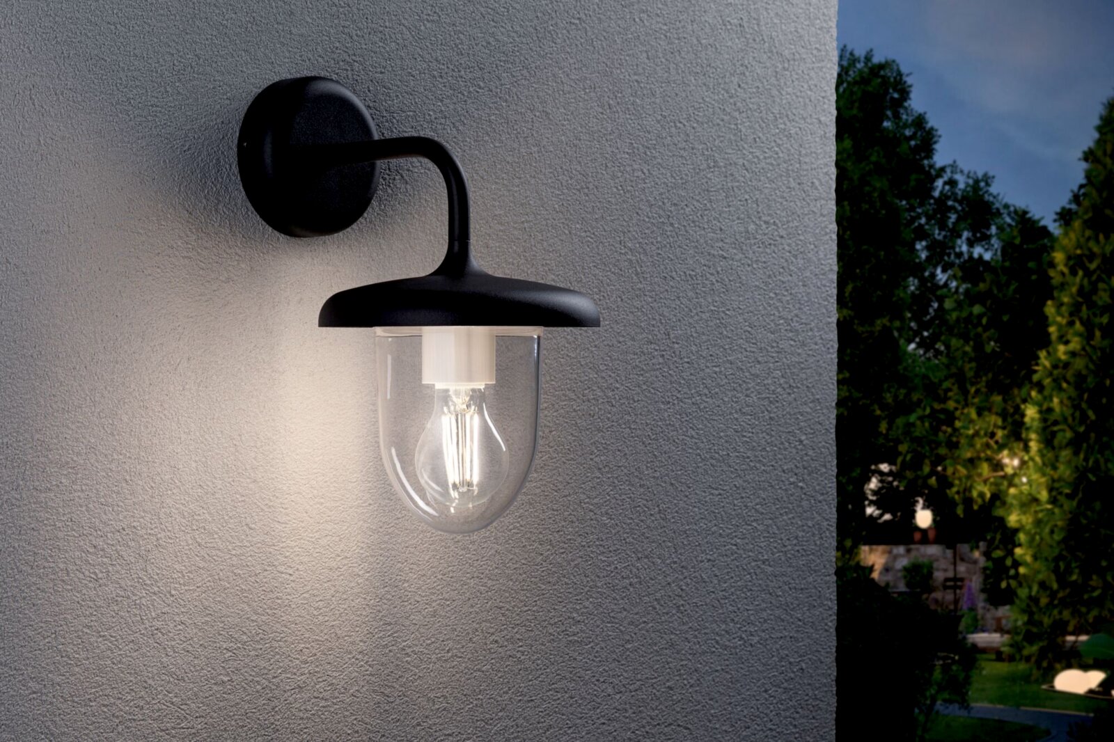 Schwarze Außen-Wandleuchte im Laternenstil mit transparentem Glas und sichtbarem Leuchtmittel an grauer Hauswand, warm leuchtend, Perspektive: leicht seitliche Frontansicht