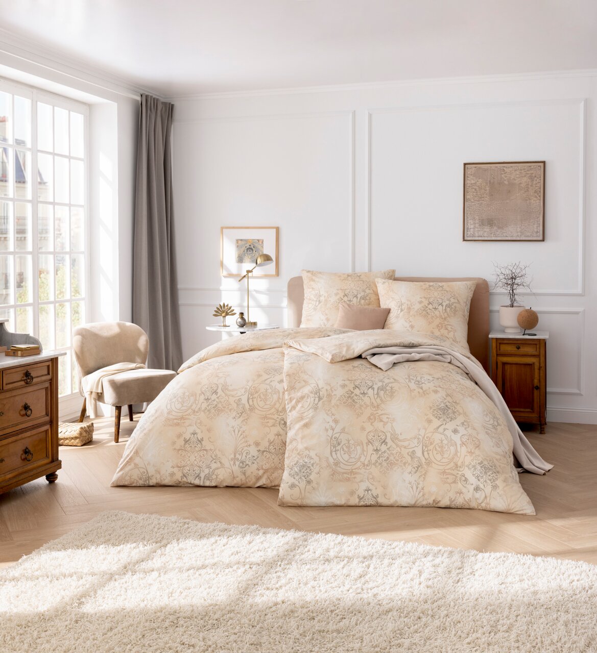 Elegant beddengoed in een licht, klassiek ontwerp met bloemmotieven, gepresenteerd in een stijlvol ingerichte slaapkamer met daglicht dat van links binnenvalt.