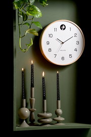 Wanduhr mit weißem Zifferblatt und schwarzem Zeigersatz, umgeben von grünen Pflanzen und schwarzen Kerzen, aus frontaler Perspektive.