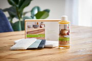 Pflege- und Reparaturset mit Flasche Renuwell Möbelöl und Reinigungstüchern auf einem Holztisch, im Hintergrund Pflanzen. Perspektive: seitlich von vorne.