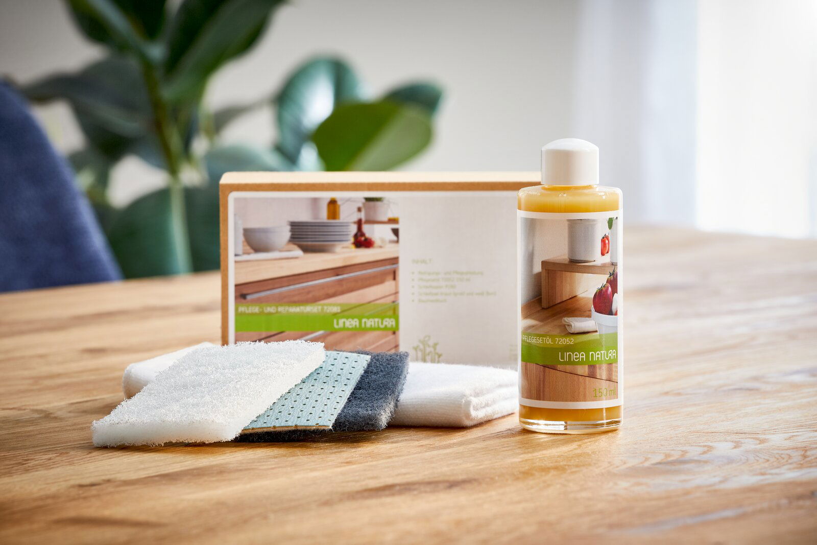 Pflege- und Reparaturset mit Flasche Renuwell Möbelöl und Reinigungstüchern auf einem Holztisch, im Hintergrund Pflanzen. Perspektive: seitlich von vorne.