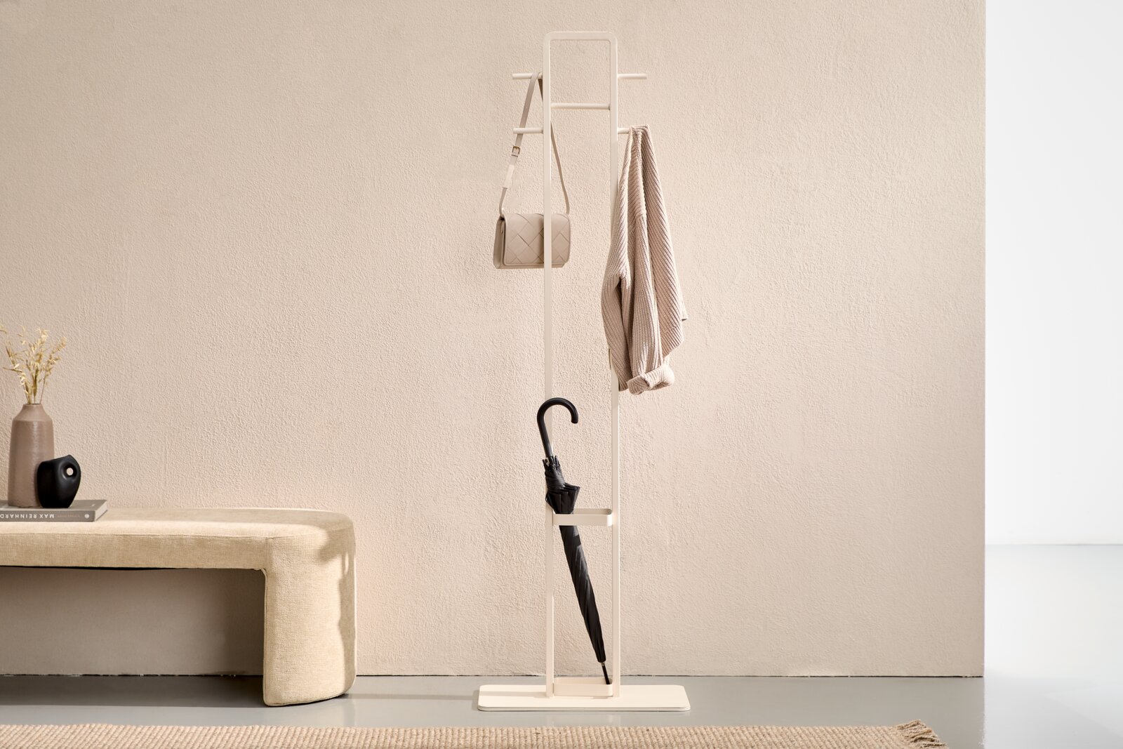 Weißer, minimalistischer Standgarderobenständer aus Metall mit Haken und Schirmhalter, mit aufgehängter Tasche und Jacke sowie eingestelltem schwarzem Regenschirm, in Frontalansicht vor heller Wand.