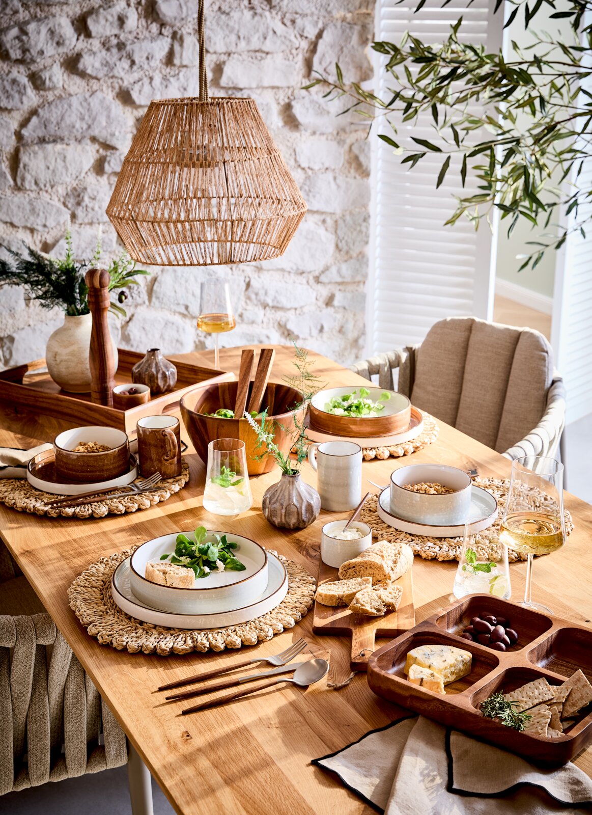 Een stijlvol gedekte eettafel met een 10-delig servies, bestaande uit borden, schalen en kopjes in natuurlijke kleuren. De tafel is van hout en versierd met gevlochten placemats, verse kruiden en een hangende lampenkap van rotan. Aanzicht van bovenaf.