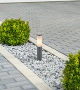 Moderne Außenleuchte BOSTON in einem Kiesbett, umgeben von grünen Büschen, aus seitlicher Perspektive fotografiert.