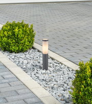 Moderne Außenleuchte BOSTON in einem Kiesbett, umgeben von grünen Büschen, aus seitlicher Perspektive fotografiert.