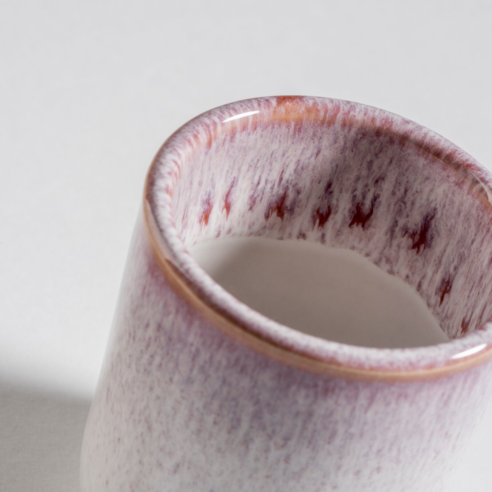 Nahaufnahme einer glasierten Keramiktasse/Becher in Weiß mit violettem Sprenkelmuster und braunem Rand, schräg von oben fotografiert