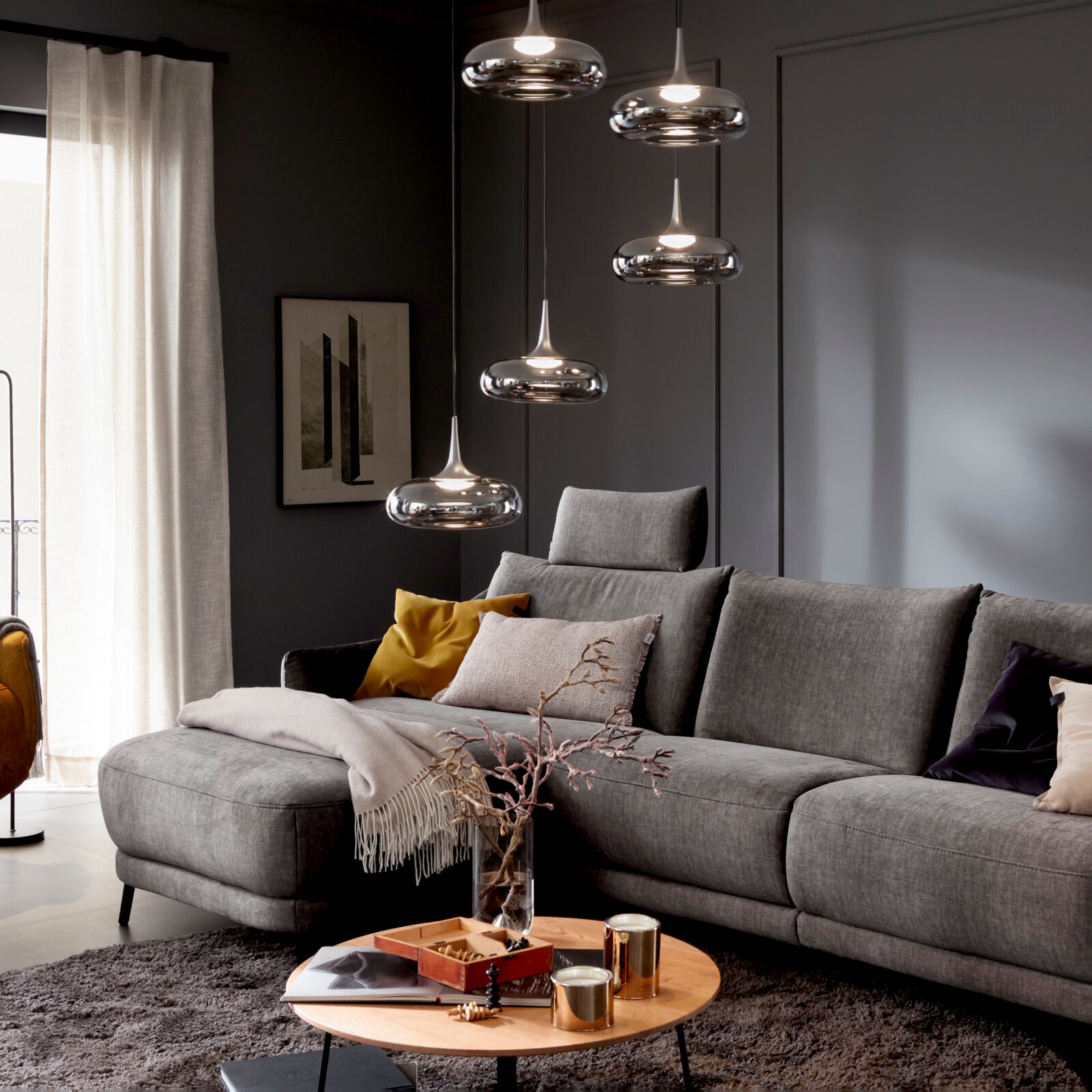 Modernes Wohnzimmer mit grauem Ecksofa und Kissen, runder Couchtisch im Vordergrund und mehreren chromfarbenen Pendelleuchten über dem Sofa; Ansicht schräg von vorn.