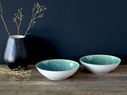 Zwei flache Keramikschalen in Weiß mit türkis-blauer Glasur innen auf rustikalem Holztisch, links eine schwarze Vase mit Zweigen vor dunkelblauem Hintergrund, Ansicht leicht von vorne