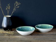 Zwei flache Keramikschalen in Weiß mit türkis-blauer Glasur innen auf rustikalem Holztisch, links eine schwarze Vase mit Zweigen vor dunkelblauem Hintergrund, Ansicht leicht von vorne
