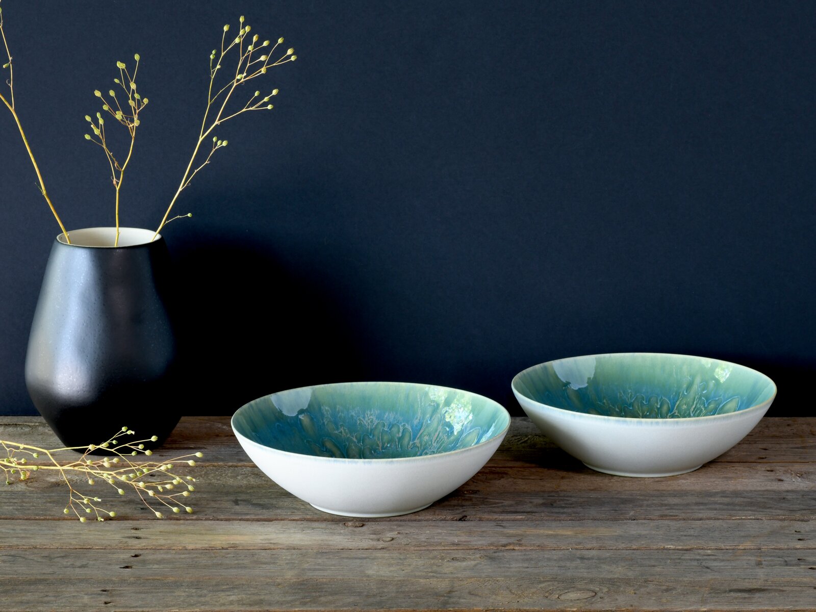 creaTable Salatschüssel 2-tlg YUKI Zwei flache Keramikschalen in Weiß mit türkis-blauer Glasur innen auf rustikalem Holztisch, links eine schwarze Vase mit Zweigen vor dunkelblauem Hintergrund, Ansicht leicht von vorne