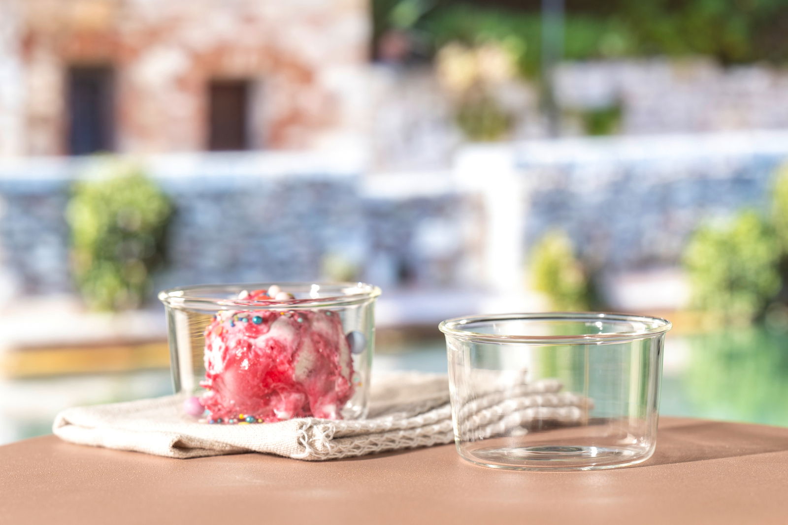 Zwei Glasschalen, eine gefüllt mit Eis und bunten Streuseln, die andere leer, auf einem Tisch mit unscharfem Hintergrund. Seitliche Perspektive.