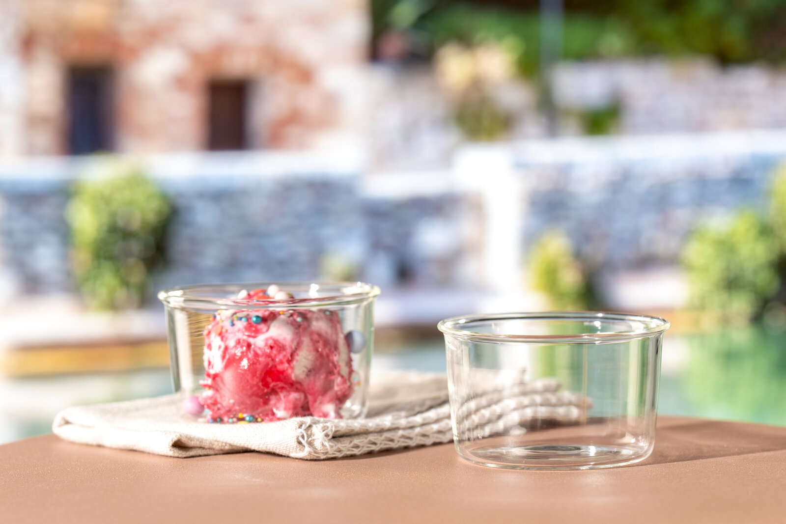 Zwei Glasschalen, eine gefüllt mit Eis und bunten Streuseln, die andere leer, auf einem Tisch mit unscharfem Hintergrund. Seitliche Perspektive.