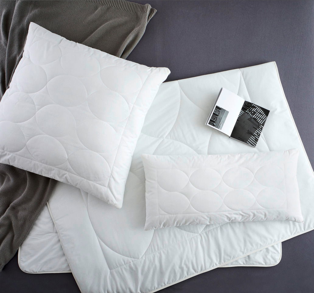 Vogelperspektive auf ein Set aus weißen Kopfkissen und Decken mit kreisförmigem Steppmuster, begleitet von einem grauen Strickstoff und einem aufgeschlagenen Buch.