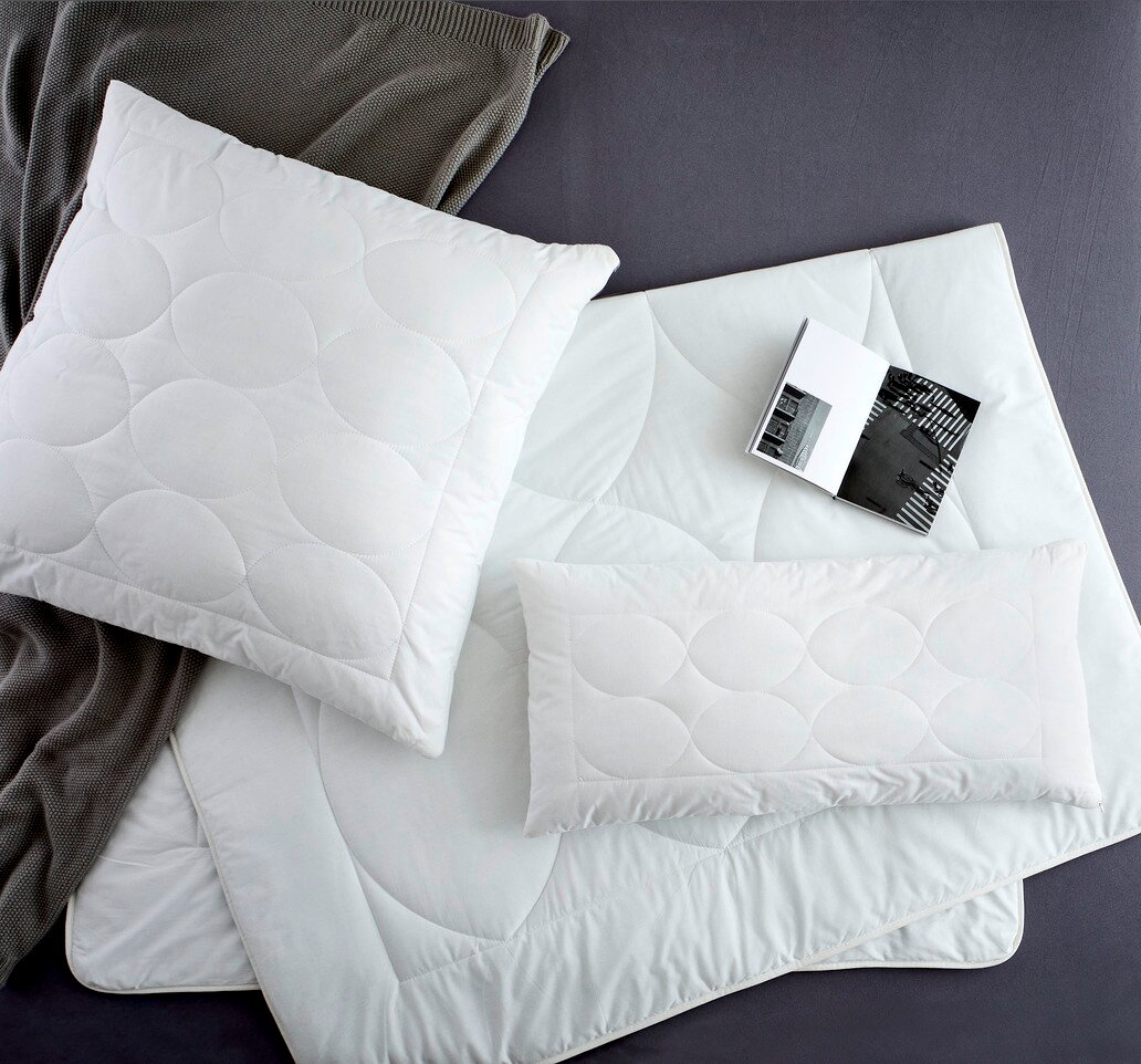 Bio-Vio Kopfkissen TENDRE Vogelperspektive auf ein Set aus weißen Kopfkissen und Decken mit kreisförmigem Steppmuster, begleitet von einem grauen Strickstoff und einem aufgeschlagenen Buch.