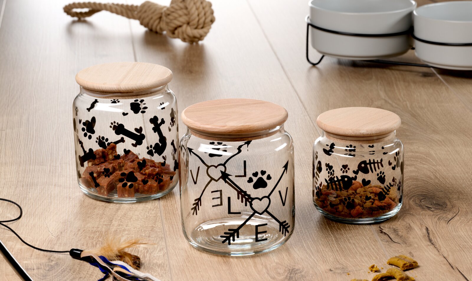 Drie glazen potten met houten deksels op een houten tafel, versierd met pootafdrukken, botten en visgraten. Op de middelste pot staat het woord 'LOVE' met pijlen. Perspectief: vooraanzicht.