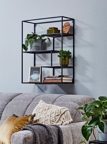Schwarzes, geometrisches Wandregal aus Metall mit mehreren Fächern, dekoriert mit Pflanzen, Büchern und Bildrahmen über einem grauen Sofa, schräg von vorn fotografiert.