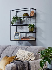 Schwarzes, geometrisches Wandregal aus Metall mit mehreren Fächern, dekoriert mit Pflanzen, Büchern und Bildrahmen über einem grauen Sofa, schräg von vorn fotografiert.