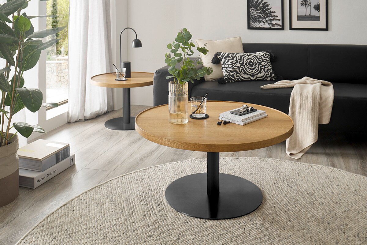 Moderner Couchtisch mit runder Holzplatte und schwarzem Metallfuß, im Wohnzimmer mit Sofa und Pflanzen, aus der Vorderperspektive.