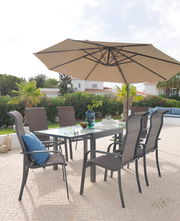 Großer, beiger Sonnenschirm über einem modernen Gartentisch mit sechs Stühlen, umgeben von grüner Vegetation und einem hellen Himmel. Perspektive von schräg unten.