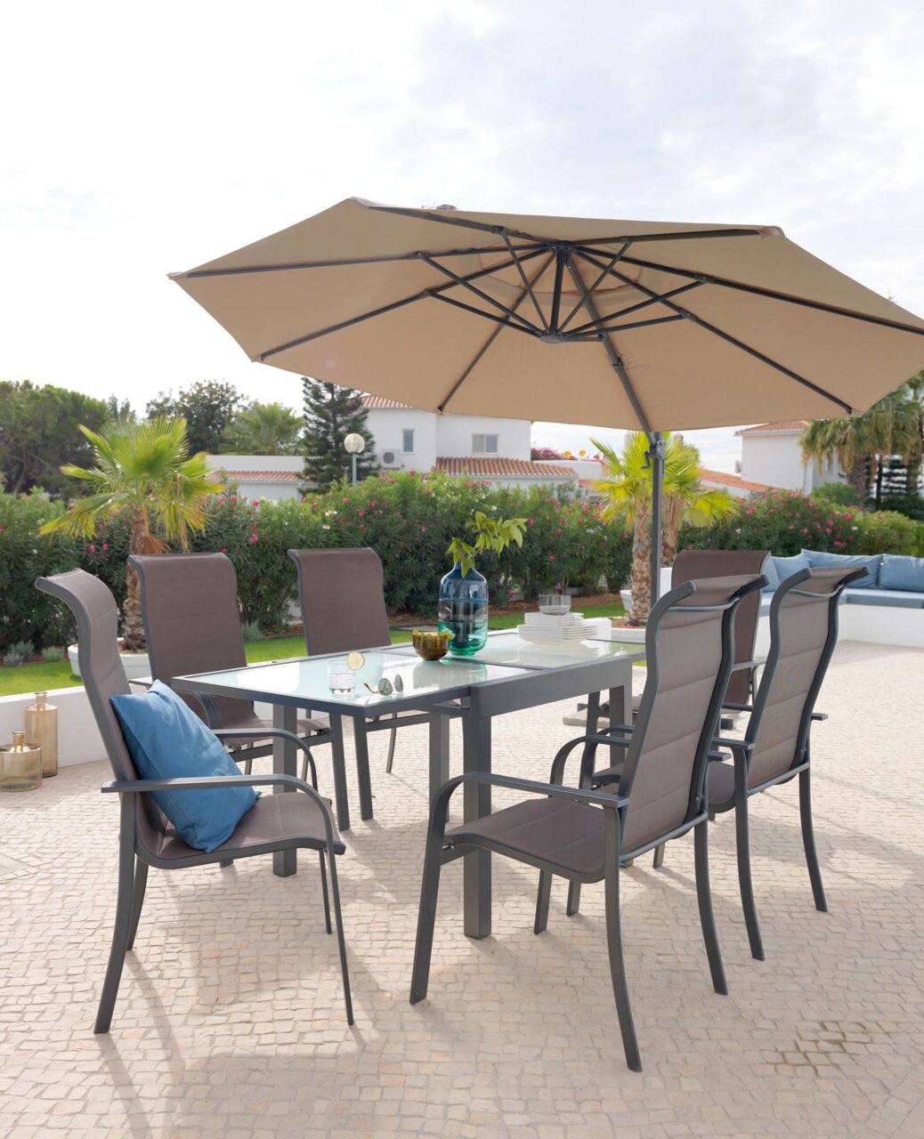 Großer, beiger Sonnenschirm über einem modernen Gartentisch mit sechs Stühlen, umgeben von grüner Vegetation und einem hellen Himmel. Perspektive von schräg unten.