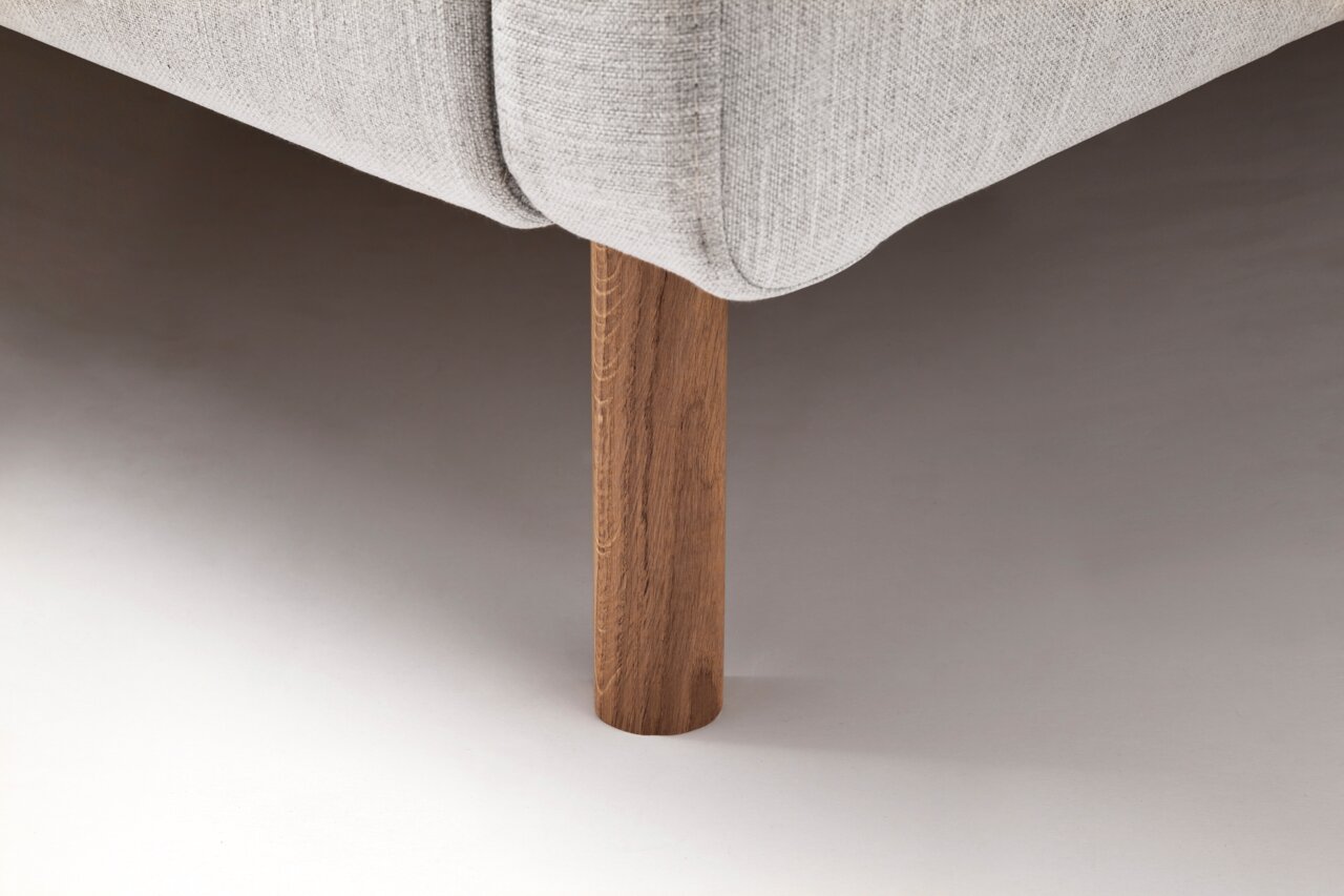 Close-up van een gestoffeerd bed met een lichtgrijze stoffen bekleding en een houten poot, gezien vanaf de zijkant.