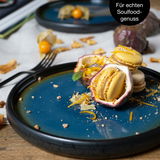 Nahaufnahme eines blauen Desserttellers mit gelben Macarons und Passionsfrucht, dekoriert mit Nüssen und Orangenschalen, auf einem Holztisch.