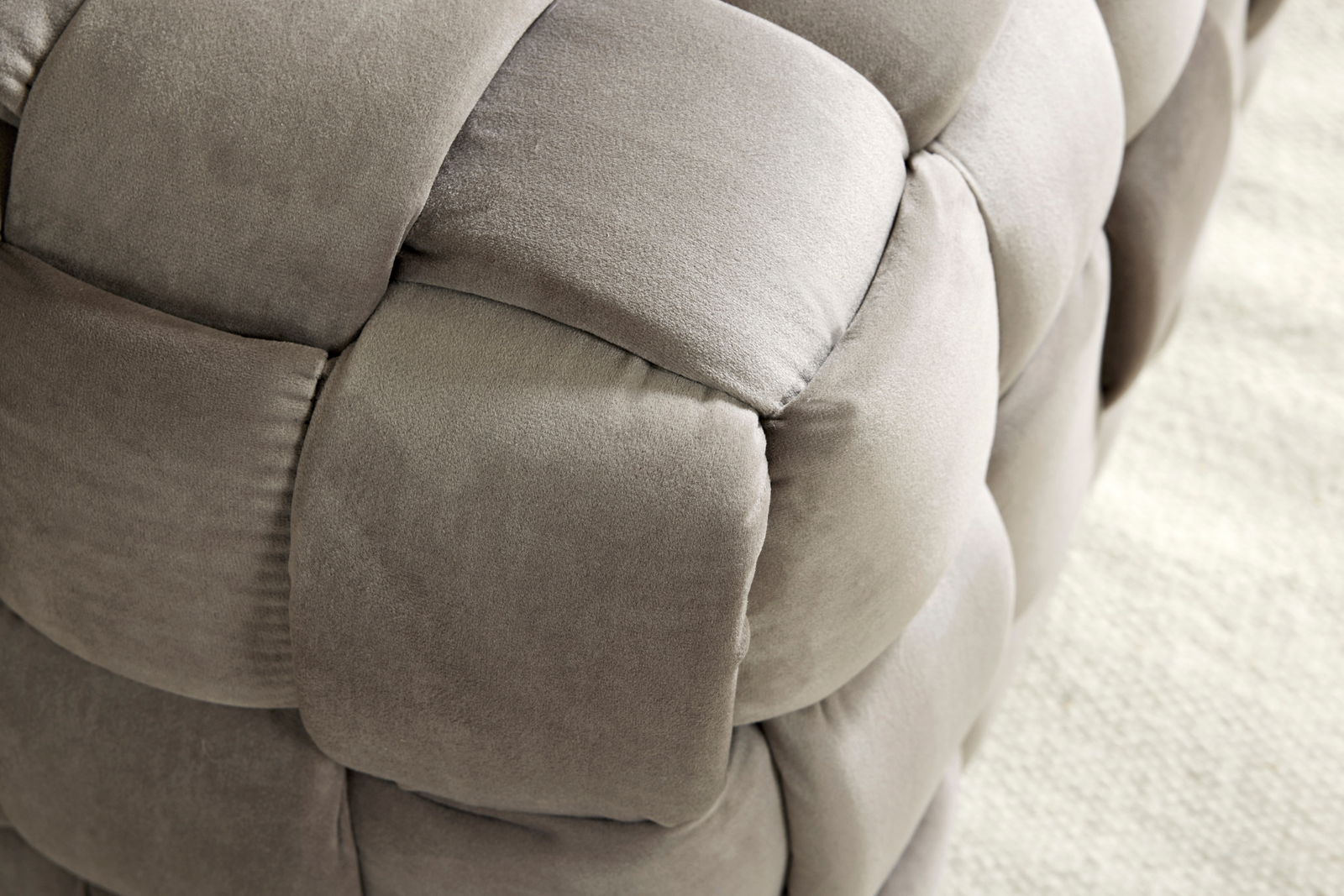 Nahaufnahme eines gepolsterten Hockers mit weichem, grauem Stoff in geflochtener Optik, seitliche Perspektive.