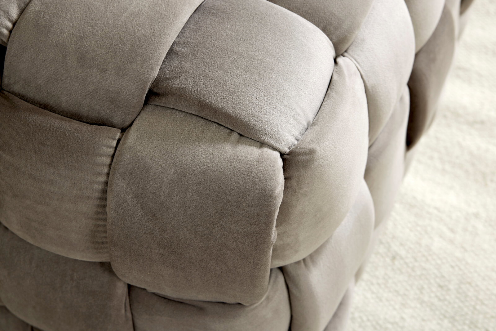 Nahaufnahme eines gepolsterten Hockers mit weichem, grauem Stoff in geflochtener Optik, seitliche Perspektive.