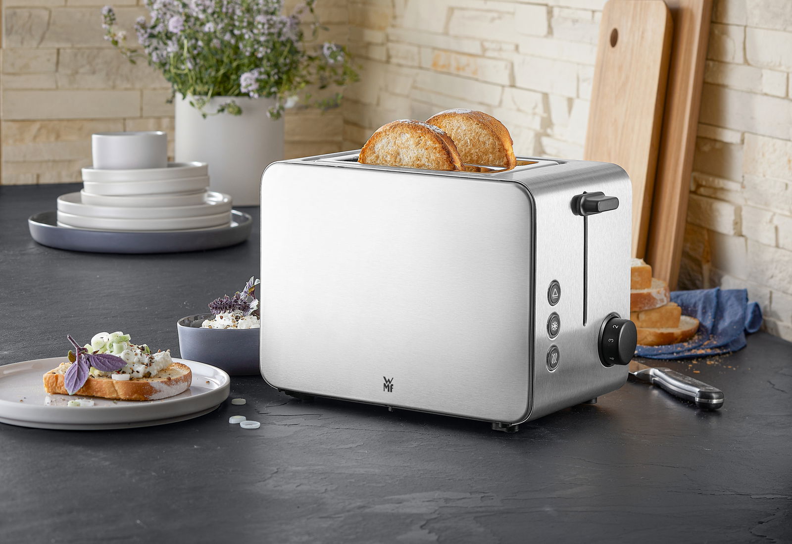 Silberner WMF Stelio Toaster Edition mit zwei Scheiben Toast, seitliche Perspektive, auf einer Küchenarbeitsplatte mit Geschirr im Hintergrund.