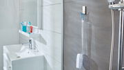 Badezimmeransicht mit tesa Powerstrips Waterproof Haken, der eine Duschbürste an einer grauen Fliesenwand hält, neben einer Dusche mit Chromarmaturen.