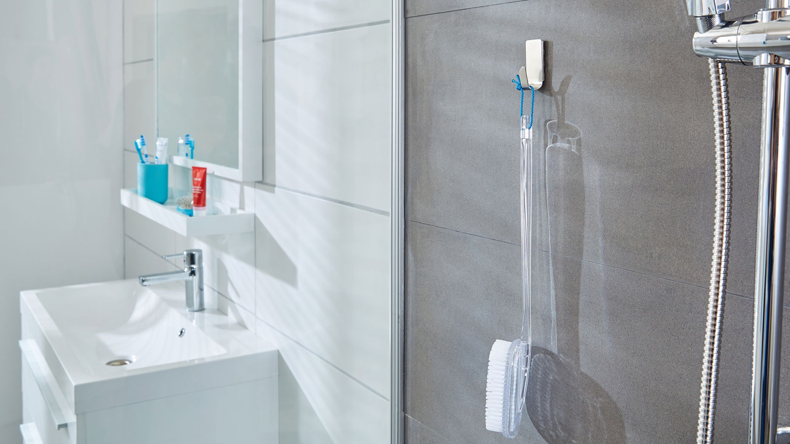 Tesa Powerstrips WATERPROOF Badezimmeransicht mit tesa Powerstrips Waterproof Haken, der eine Duschbürste an einer grauen Fliesenwand hält, neben einer Dusche mit Chromarmaturen.