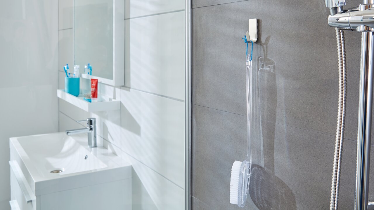 Tesa Powerstrips WATERPROOF Badezimmeransicht mit tesa Powerstrips Waterproof Haken, der eine Duschbürste an einer grauen Fliesenwand hält, neben einer Dusche mit Chromarmaturen.