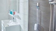 Badezimmeransicht mit tesa Powerstrips Waterproof Haken, der eine Duschbürste an einer grauen Fliesenwand hält, neben einer Dusche mit Chromarmaturen.