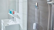 Badezimmeransicht mit tesa Powerstrips Waterproof Haken, der eine Duschbürste an einer grauen Fliesenwand hält, neben einer Dusche mit Chromarmaturen.