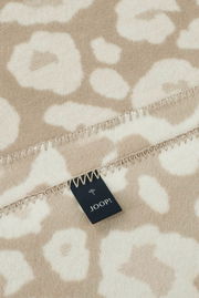 Nahaufnahme einer beige-weißen Wohndecke mit floralem Muster und einem Joop-Label, perspektivisch von oben fotografiert.