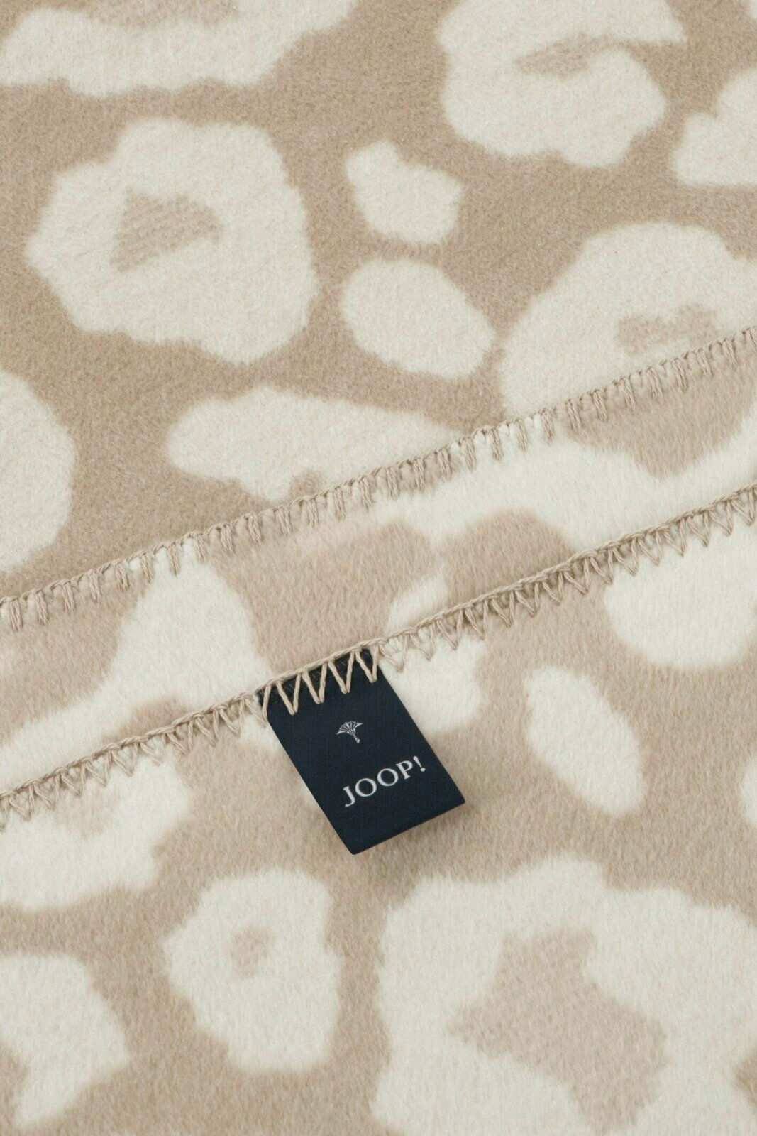 Nahaufnahme einer beige-weißen Wohndecke mit floralem Muster und einem Joop-Label, perspektivisch von oben fotografiert.