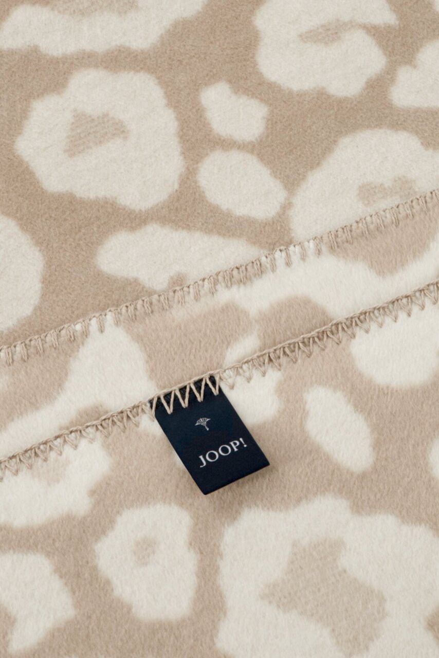 Close-up van een beige-witte woondeken met bloemmotief en een Joop-label, gefotografeerd vanuit een perspectief van bovenaf.