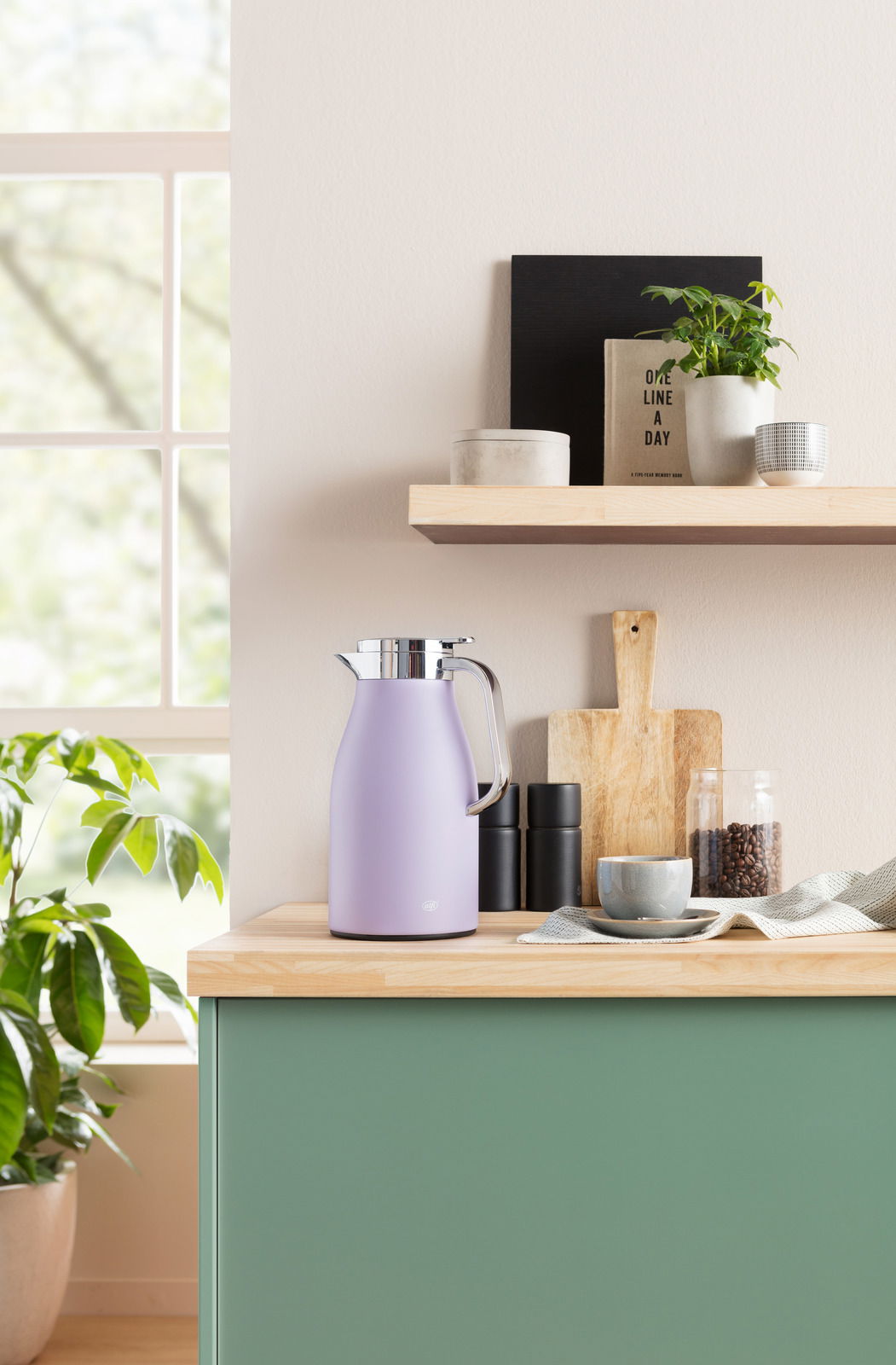 Lila Isolierkanne Skyline auf einer Küchenarbeitsplatte, seitliche Perspektive, umgeben von Küchenutensilien und Pflanzen.
