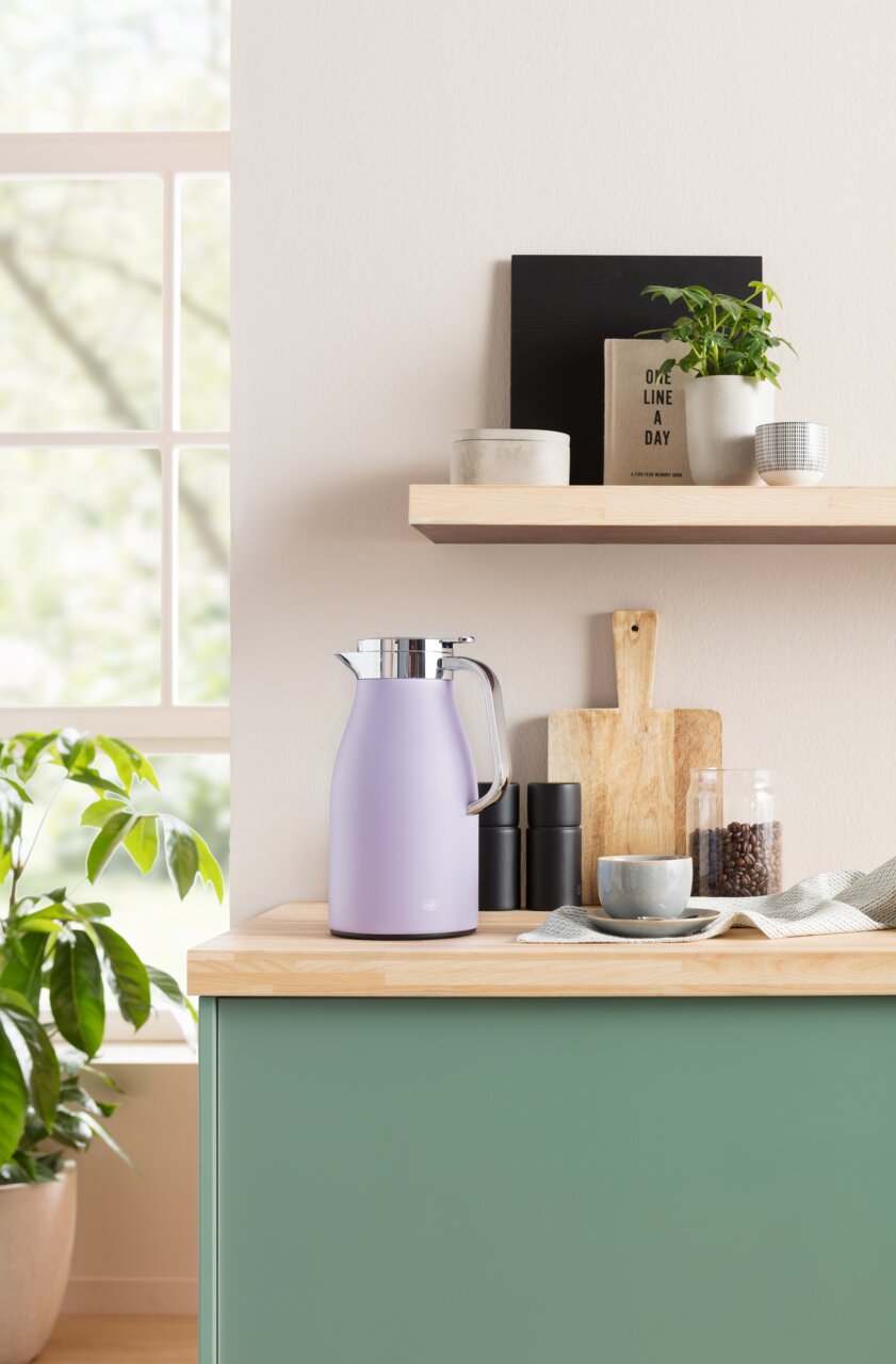 Lila Isolierkanne Skyline auf einer Küchenarbeitsplatte, seitliche Perspektive, umgeben von Küchenutensilien und Pflanzen.