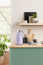 Lila Isolierkanne Skyline auf einer Küchenarbeitsplatte, seitliche Perspektive, umgeben von Küchenutensilien und Pflanzen.