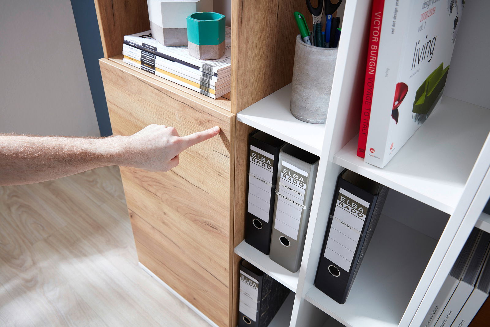 Nahaufnahme eines Aktenschranks mit Holz- und weißen Elementen, gefüllt mit Ordnern und Büchern. Eine Hand zeigt auf die Holzverkleidung. Perspektive von schräg oben.