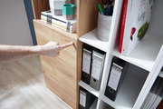 Nahaufnahme eines Aktenschranks mit Holz- und weißen Elementen, gefüllt mit Ordnern und Büchern. Eine Hand zeigt auf die Holzverkleidung. Perspektive von schräg oben.