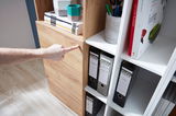 Nahaufnahme eines Aktenschranks mit Holz- und weißen Elementen, gefüllt mit Ordnern und Büchern. Eine Hand zeigt auf die Holzverkleidung. Perspektive von schräg oben.