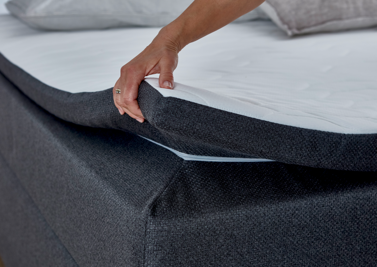 Nahaufnahme eines Boxspringbetts mit einer Hand, die die Matratze anhebt, um die Struktur und den Bezug zu zeigen.