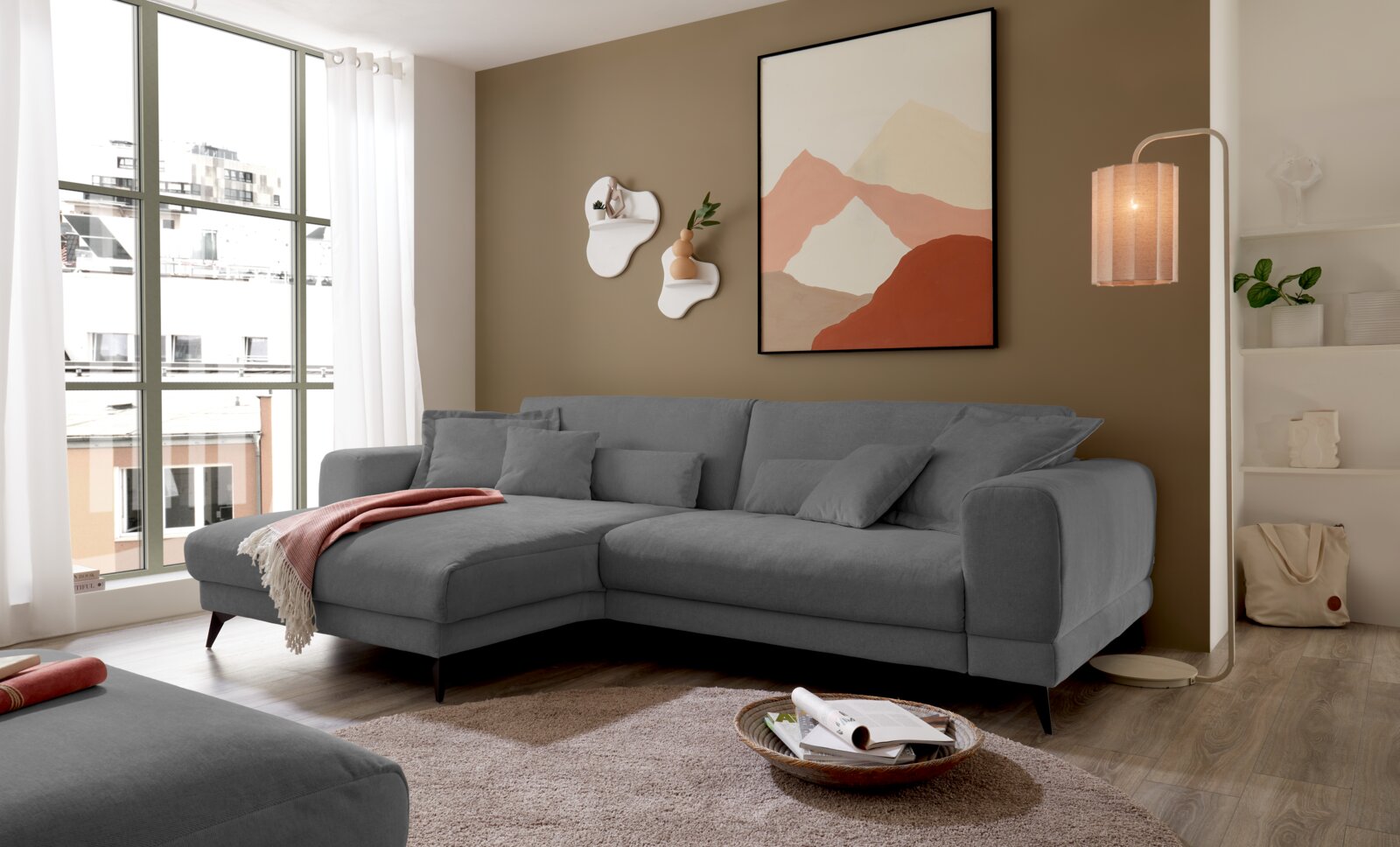Set one by Musterring Ecksofa SET ONE MIDLAND Modernes graues L-förmiges Ecksofa mit Recamiere links und mehreren Kissen im hellen Wohnzimmer; Ansicht schräg von vorne (3/4-Perspektive) mit Fenster links und Stehlampe rechts.