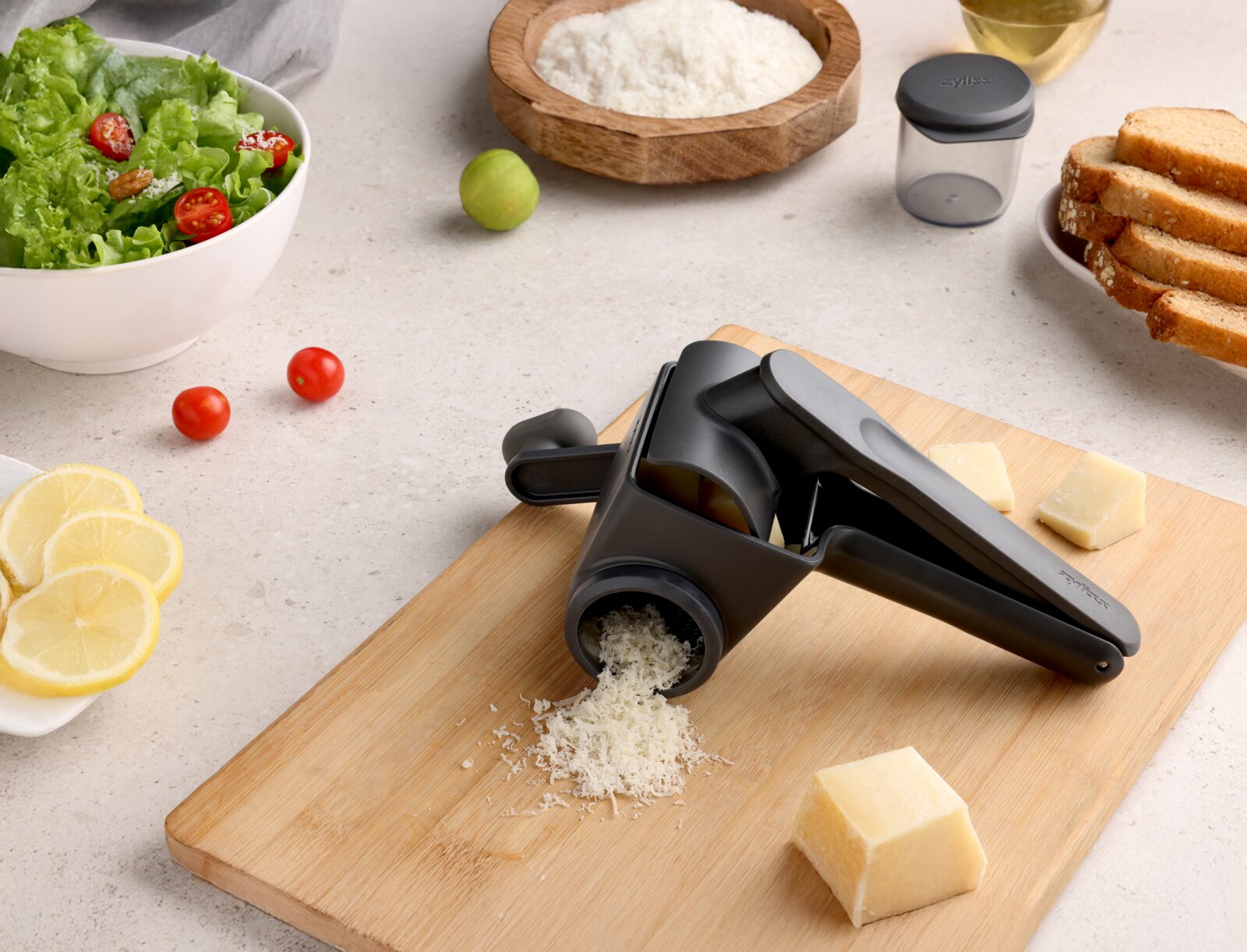 Schwarze Handkurbel-Reibe für Käse auf Holzschneidebrett mit frisch geriebenem Käse und Parmesanwürfeln, aufgenommen aus schräger Draufsicht.