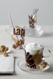 Latte Macchiato Löffel in einem Glas mit Schriftzug, von vorne fotografiert, mit einem weiteren Glas im Hintergrund.