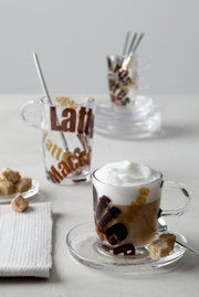 Latte Macchiato Löffel in einem Glas mit Schriftzug, von vorne fotografiert, mit einem weiteren Glas im Hintergrund.