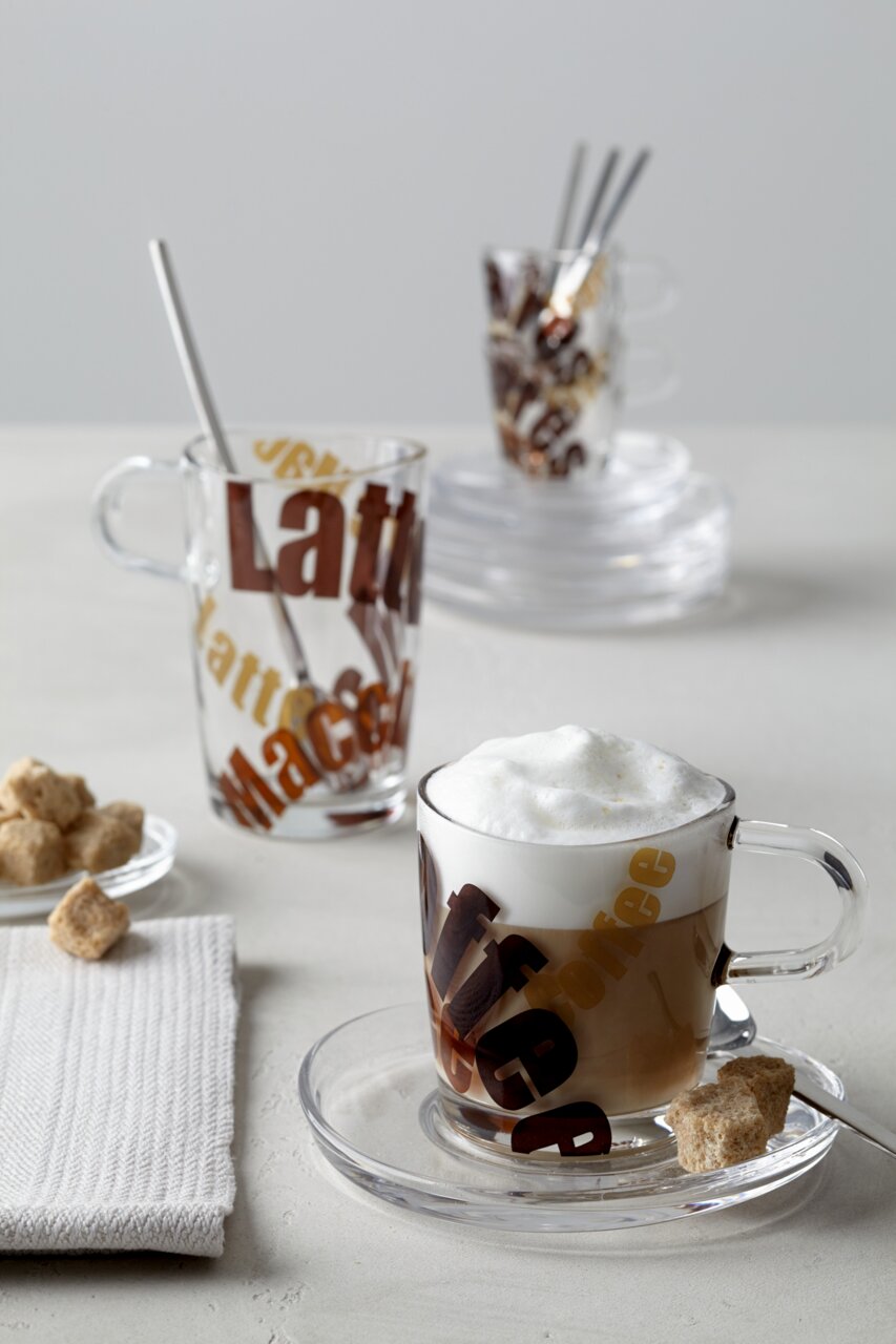 Een latte macchiato-lepel in een glas met opschrift, gefotografeerd van voren, met nog een glas op de achtergrond.